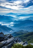 Majestic mountain ranges layered in misty blue hues stretch to horizon under dramatic cloudy sky, with rocky cliff overlook in foreground creating depth perspective.