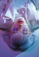 Young woman with long hair wearing futuristic sunglasses and chain necklace, lying down in purple and blue neon lighting. Surreal fashion and beauty concept for modern design projects.