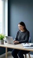 Young professional woman working on laptop at modern office desk with plant and natural light from window in contemporary workspace.