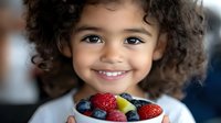 Mixed race child with curly hair holding bowl of fresh berries, expressing joy and healthy eating habits with natural bright smile against blurred background.