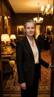 Professional mature Caucasian businesswoman in elegant black suit standing confidently in luxury hotel lobby with warm lighting and classic decor.