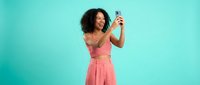Young African American woman in pink outfit taking selfie with smartphone against turquoise background, smiling happily while posing for social media photo.