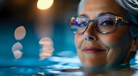 Mature woman with stylish glasses and silver hair enjoying evening swim in pool with bokeh lights creating dreamy atmosphere. Wellness and active aging concept.