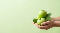 Fresh green apples with leaves held in open hands against pale background, promoting healthy eating and organic produce for wellness campaigns.