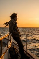 Young woman with flowing hair standing on boat deck during golden sunset over ocean waves enjoying maritime adventure and sea breeze freedom.
