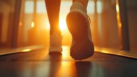 Close-up view of running shoes on treadmill during sunrise workout with golden sunlight streaming through windows, creating dramatic silhouette and lens flare effect.