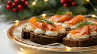 Festive smoked salmon canapes with cream cheese and dill on dark bread, served on elegant plate with Christmas tree and lights in background for holiday entertaining.
