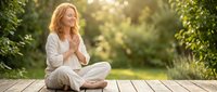 Middle-aged Caucasian woman in white clothing sitting cross-legged on wooden deck practicing meditation outdoors surrounded by lush green nature during golden hour.