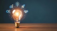 Glowing light bulb with business innovation icons floating around it on wooden surface against dark background for creative startup concepts.