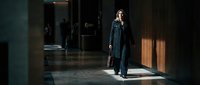 Professional businesswoman in dark suit walking through modern office corridor with dramatic lighting and shadows creating corporate atmosphere.