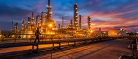 Industrial oil refinery complex with illuminated towers and processing units against dramatic sunset sky creating golden hour atmosphere for energy sector.