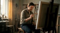 Middle-aged Caucasian male artist painting on canvas in sunlit studio workspace with easel and art supplies for creative professional content.