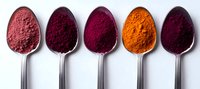 Vibrant spices and natural food coloring powders displayed in silver spoons, ranging from pink to orange and deep purple hues, arranged in minimalist composition.