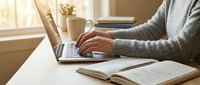 Person in a gray sweater typing on a laptop at a wooden desk with an open notebook, pen, and coffee mug in warm natural light.