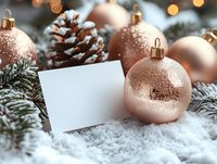 Rose gold Christmas baubles with blank white card on snowy pine branches and pinecones, festive bokeh lights in background create magical holiday atmosphere.