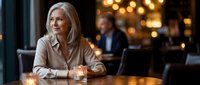 Mature blonde businesswoman sitting at restaurant table with glass, contemplating during evening meeting with colleague in background under warm lighting.