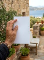 Male hand holding blank white paper against Mediterranean coastal villa backdrop with stone architecture, olive trees, and sea view, suitable for mockups, advertising, and promotional campaigns.