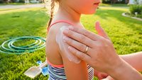 Caucasian child with blonde hair receiving sunscreen application on shoulder outdoors in sunny backyard setting with hula hoop, promoting sun protection and summer safety for families.