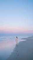 Young woman walking alone on peaceful beach at sunset with pink and blue sky colors reflecting on wet sand and gentle waves.