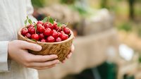 Fresh red cherries in wooden bowl held by person in white sleeve, garden setting with blurred background.