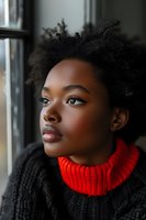 Young African American woman with natural afro hair gazing thoughtfully through window, wearing black sweater with striking red turtleneck, captured in soft natural lighting.