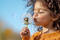 Mixed race curly haired child blowing dandelion seeds in spring garden. Close up portrait of young girl in orange sweater making wish against blue sky background.