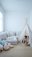 Two young children playing inside white teepee tent in modern minimalist playroom with gray sofa and colorful stuffed animals on wooden floor.