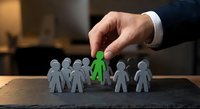Businessman hand selecting green paper figure from group of people cutouts on wooden desk for recruitment and talent acquisition in corporate office.