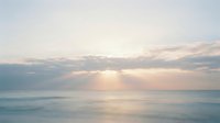 Serene ocean sunrise with soft golden light breaking through clouds over calm waters, creating peaceful atmosphere for wellness and nature backgrounds.