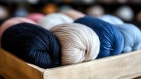 Colorful yarn skeins in navy blue, white and denim blue shades arranged in wooden box. Close-up view of soft merino wool balls with selective focus.