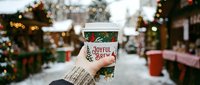 Gloved hand holding festive holiday coffee cup with joyful brew text in snowy winter street market setting with decorated trees and lights.