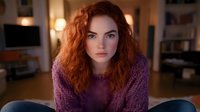 Young redhead woman with curly hair wearing purple sweater sitting indoors looking at camera with serious expression in cozy home environment.