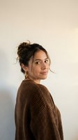 Young Caucasian woman with messy bun hairstyle wearing brown knit sweater looking over shoulder with gentle smile against neutral beige wall background.