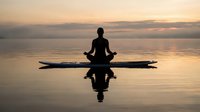 Silhouette of person meditating in lotus position on paddleboard at sunset over calm water with golden sky reflection creating peaceful wellness scene.
