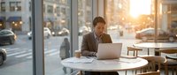 Professional businessman working on laptop in modern office cafe during golden hour sunset with city street view through large windows.