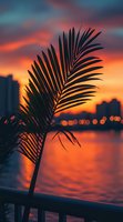 Silhouette of palm frond against vibrant orange and pink sunset sky over city waterfront, with bokeh lights reflecting on calm water surface. Perfect for travel content.