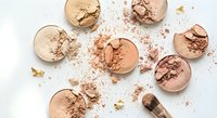 Crushed face powder makeup compacts in nude and bronze shades scattered on white background with cosmetic brush for beauty and fashion applications.