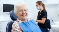 Happy elderly Caucasian woman patient smiling in dental chair with protective bib while female dentist prepares treatment in modern clinic background.