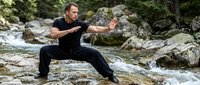 Middle-aged Caucasian man practicing martial arts stance on rocky riverbank with flowing water and boulders, demonstrating balance and strength training outdoors.
