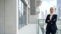 Middle Eastern woman in professional business attire standing confidently on modern office balcony with city skyline view, arms crossed in power pose.