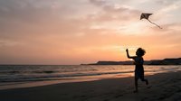 Silhouetted child flying kite on sandy beach at sunset with ocean waves and golden sky, outdoor recreation and leisure activity.