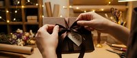 Hands tying a satin ribbon bow on a kraft paper gift box with a love tag in a cozy decorated workshop with warm lights.