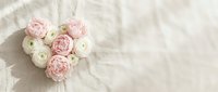 Heart-shaped arrangement of pink and white ranunculus flowers on soft beige textured background for romantic wedding or Valentine's Day celebrations.