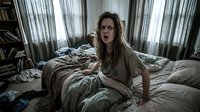Caucasian woman with long brown hair and glasses, appearing shocked or surprised, sitting on unmade bed surrounded by scattered clothing and clutter in bedroom with sheer curtains, conveying stress or overwhelm.