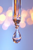 Crystal clear water drop with golden light reflections suspended against soft bokeh background in pastel blue and pink tones, macro photography.
