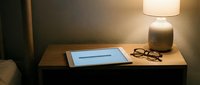 Cozy bedroom nightstand with tablet, glasses, and glowing lamp in soft evening light, creating a relaxing bedtime reading atmosphere.
