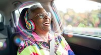 Happy senior African American woman enjoying music with colorful headphones while traveling in car backseat during sunny day road trip adventure.