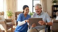 Asian female healthcare worker in blue scrubs showing tablet to smiling elderly Caucasian man in home care setting for medical consultation and support.