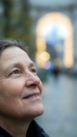 Mature woman with light skin tone gazing upward thoughtfully in urban setting with blurred architectural background, conveying contemplation and hope.