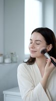 Young Asian woman applying makeup with brush in modern bathroom wearing white robe. Morning beauty routine skincare cosmetics self-care wellness lifestyle.